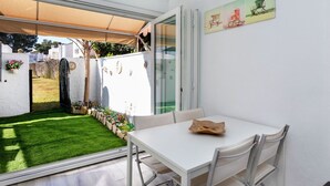 Een moderne patio met een witte tafel en stoelen, een glazen deur naar een tuin en een aan de muur bevestigd schilderij.