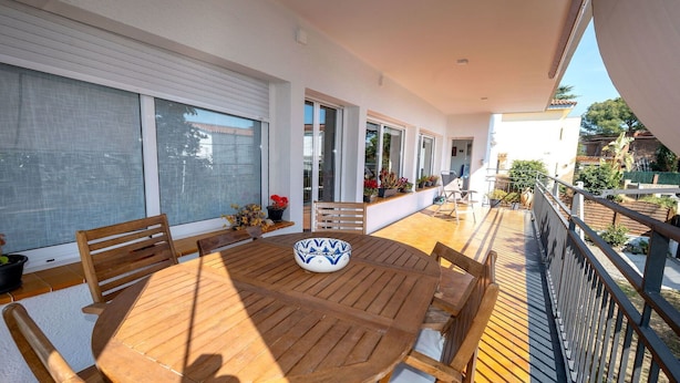 Ein Holztisch mit Stühlen auf einem Balkon, der durch eine Brüstung gesichert ist. Übertöpfe mit Pflanzen und Blick auf ein Gebäude.