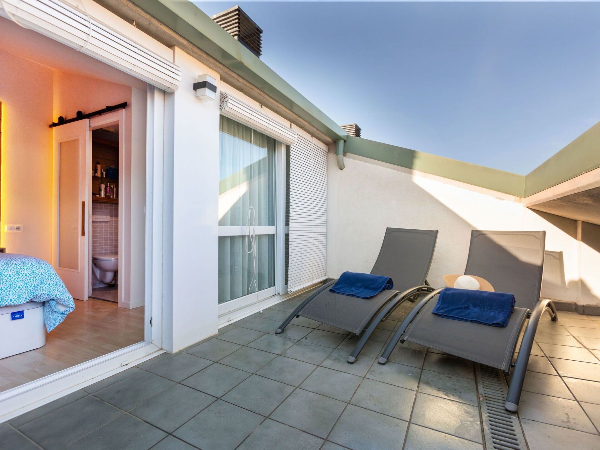 A balcony with two sun loungers, a door leading to a bathroom, and a tiled floor.