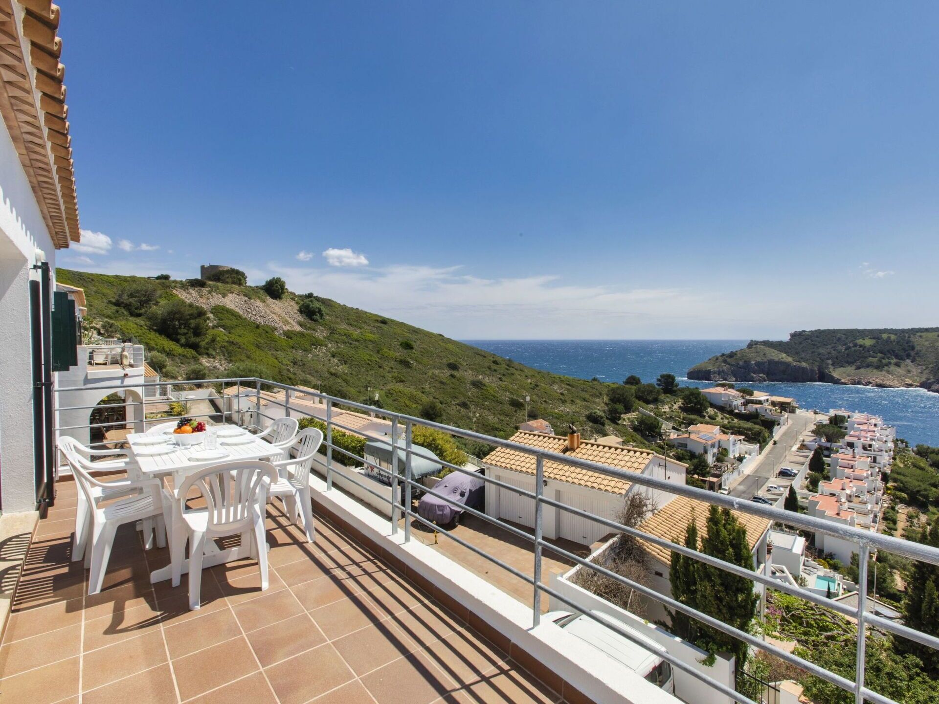 A balcony with a table and chairs, overlooking a coastal town and the sea.