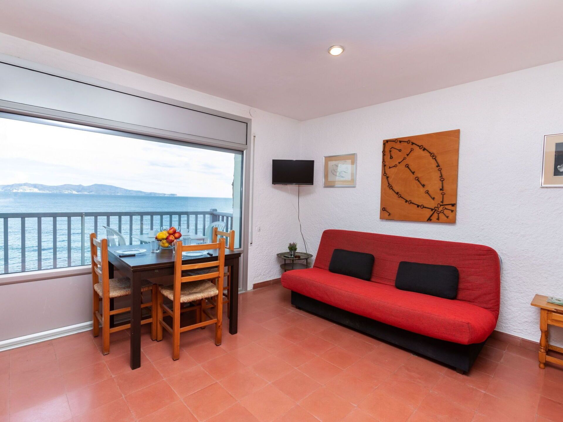 A room with a dining table and chairs, a red sofa, a TV, and a large window with a view of the sea.