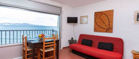 A room with a dining table and chairs, a red sofa, a TV, and a large window with a view of the sea.