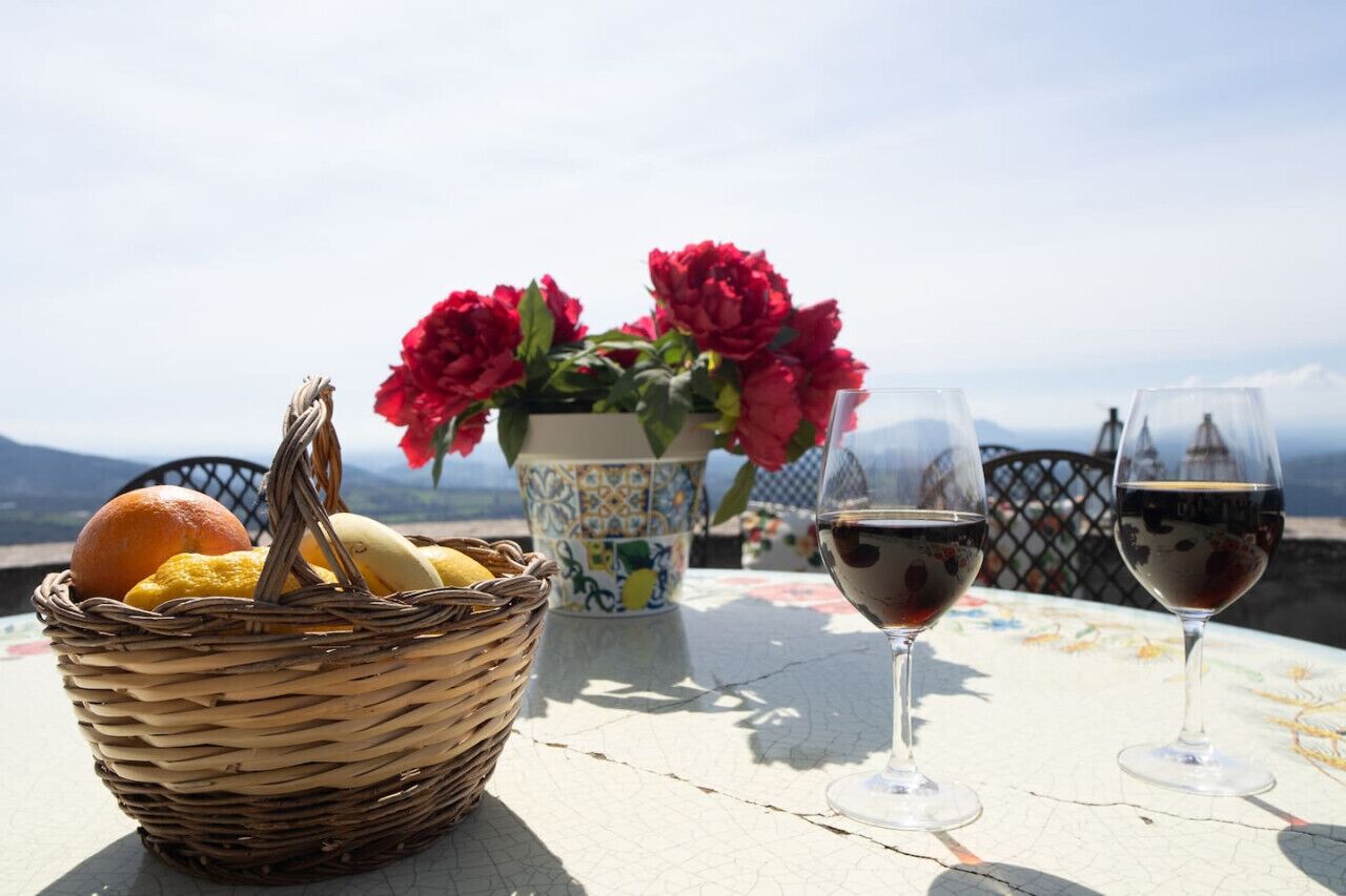 Una mesa con una cesta de mimbre que contiene pan y fruta, un florero con flores rojas y dos copas de vino sobre una mesa con vistas a las montañas.