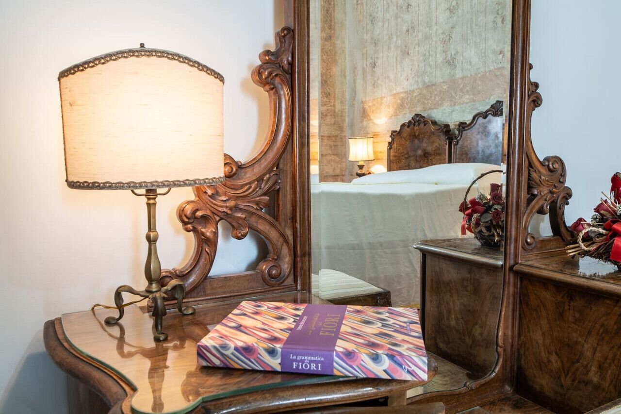 A wooden bedside table with a lamp, a mirror, and a book titled "La grammatica Fiori".