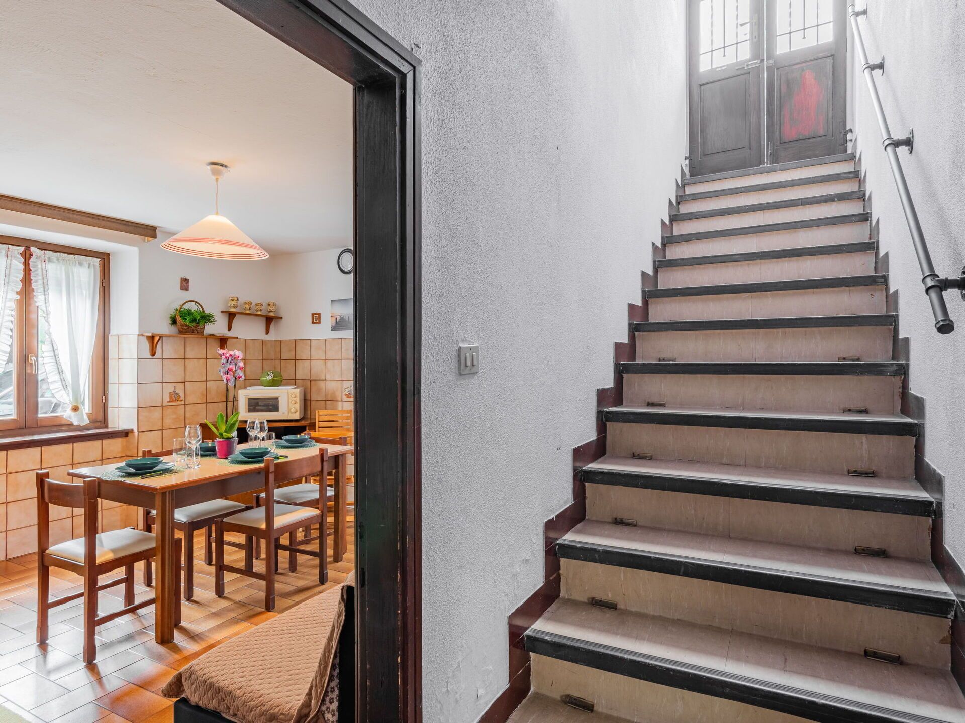 A staircase with a handrail, leading to a room with a dining area, a microwave, and a window with curtains.