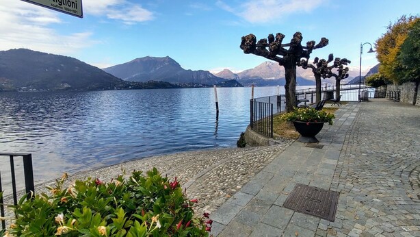 A waterfront promenade with a sign reading "Lungolago Castiglioni," a stone pathway, a metal fence, and a row of trees with hanging ornaments.