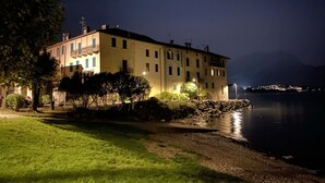 A large, multi-story building with a balcony on the second floor, situated by a lake with a rocky shoreline.