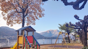 A playground with a slide, climbing steps, and swings, set against a backdrop of mountains and a lake.