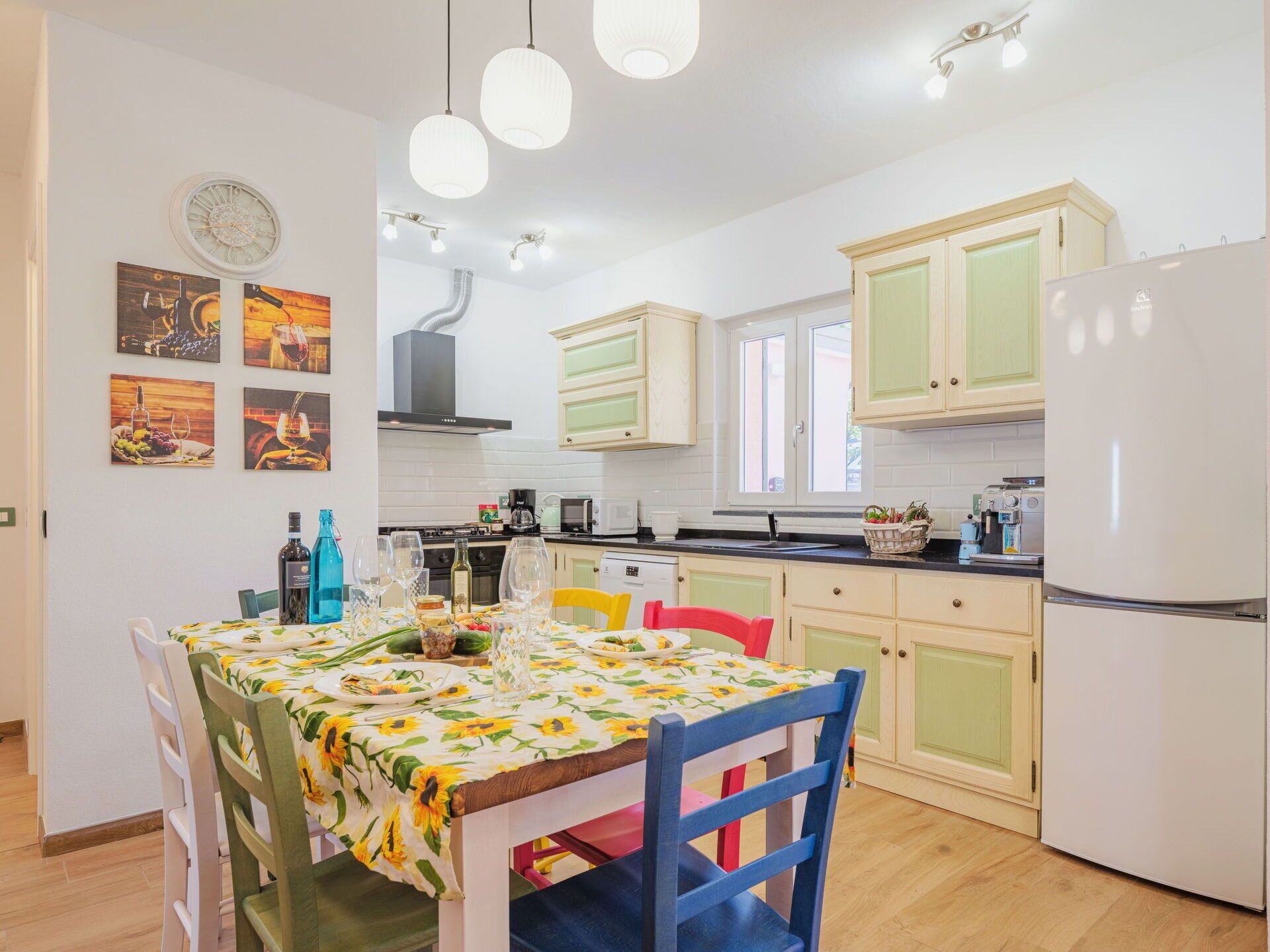 A modern kitchen with light green cabinets, a white refrigerator, and a dining table set with a floral tablecloth.