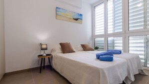 A bedroom with two beds, a nightstand with a lamp, a painting on the wall, and large windows with blinds.