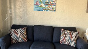 A living room with a dark blue sofa, two patterned throw pillows, and a white coffee table. A colorful painting of a coastal scene hangs on the wall.