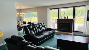 Een woonkamer met een zwarte leren bank, een houten salontafel en een open haard. Door grote ramen is er een eetgedeelte zichtbaar met een tafel en stoelen.