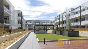 A modern apartment complex with multiple balconies, a central courtyard with artificial grass, and a clear sky.
