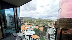 Un balcon avec deux fauteuils en osier et une petite table, d’où l’on peut admirer un paysage urbain avec des bâtiments et une colline.