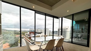 Une pièce moderne avec une table et des chaises en verre, de grandes fenêtres donnant sur la ville et un sol recouvert de moquette.