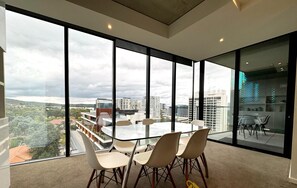 Een moderne kamer met een glazen tafel en stoelen, grote ramen met uitzicht over de stad, en een vloerbedekking.
