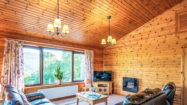 A cozy cabin interior with wooden walls, a large window with curtains, a sofa, a coffee table, a TV, and a fireplace. - Nice apartment in villa for 6 guests with WIFI, TV, patio and panoramic view (Inverness)