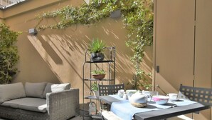 Un espace patio aménagé avec un canapé en osier, une table dressée avec de la vaisselle blanche et une étagère en métal ornée de plantes.