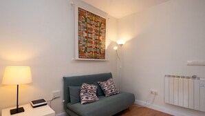 Ett litet, ombonat rum med en grå soffa, ett vitt sidobord med en lampa och en färgstark, abstrakt tavla på väggen.