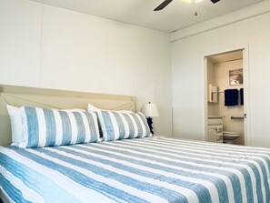 A neatly made bed with striped bedding, a bedside lamp, and a view of a bathroom with a toilet and towel rack.