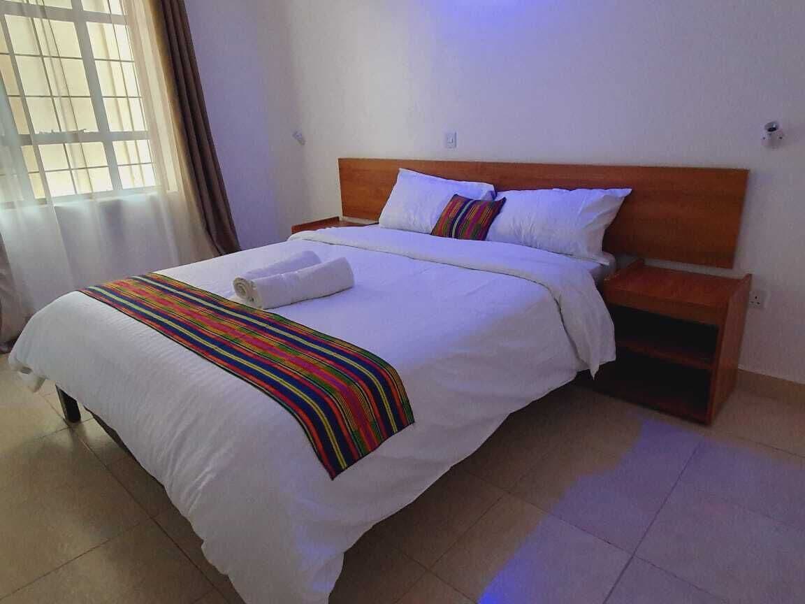 Una cama bien tendida con sábanas blancas y un colorido edredón a rayas, un cabecero de madera y una mesita de noche con una lámpara.