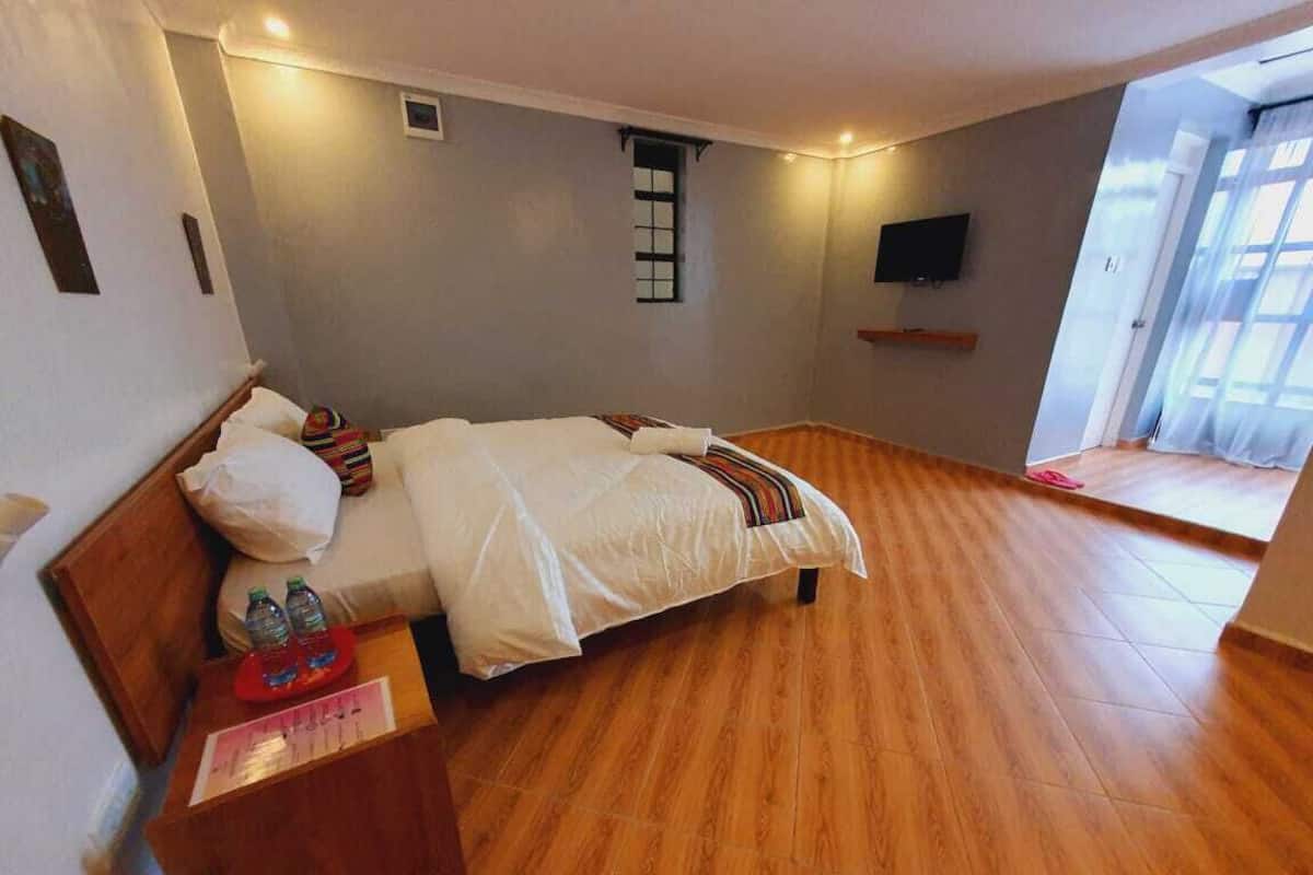 Quarto com piso de madeira, cama com roupa de cama branca, mesinha de cabeceira pequena com bandeja vermelha, televisão montada na parede e janela com cortinas.