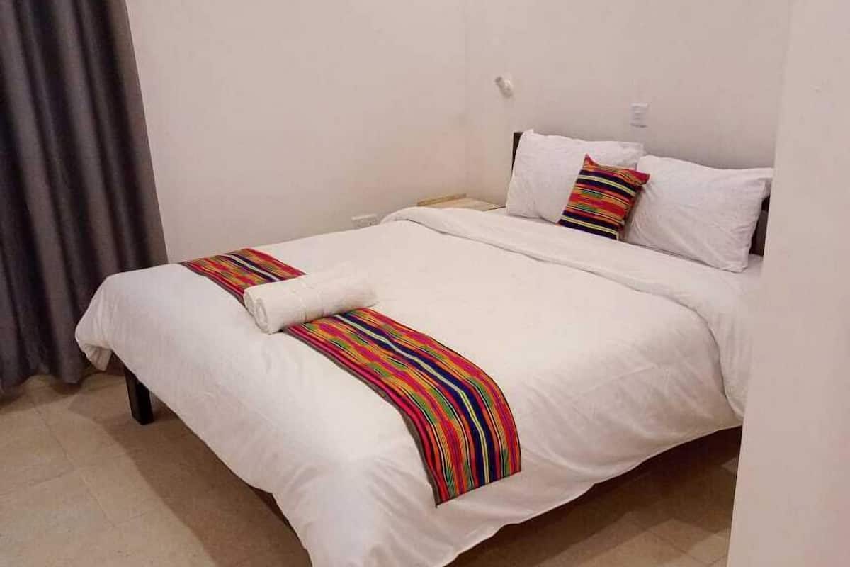 Uma cama de solteiro com roupa de cama branca e almofadas coloridas listradas, cortina cinza e chão ladrilhado.