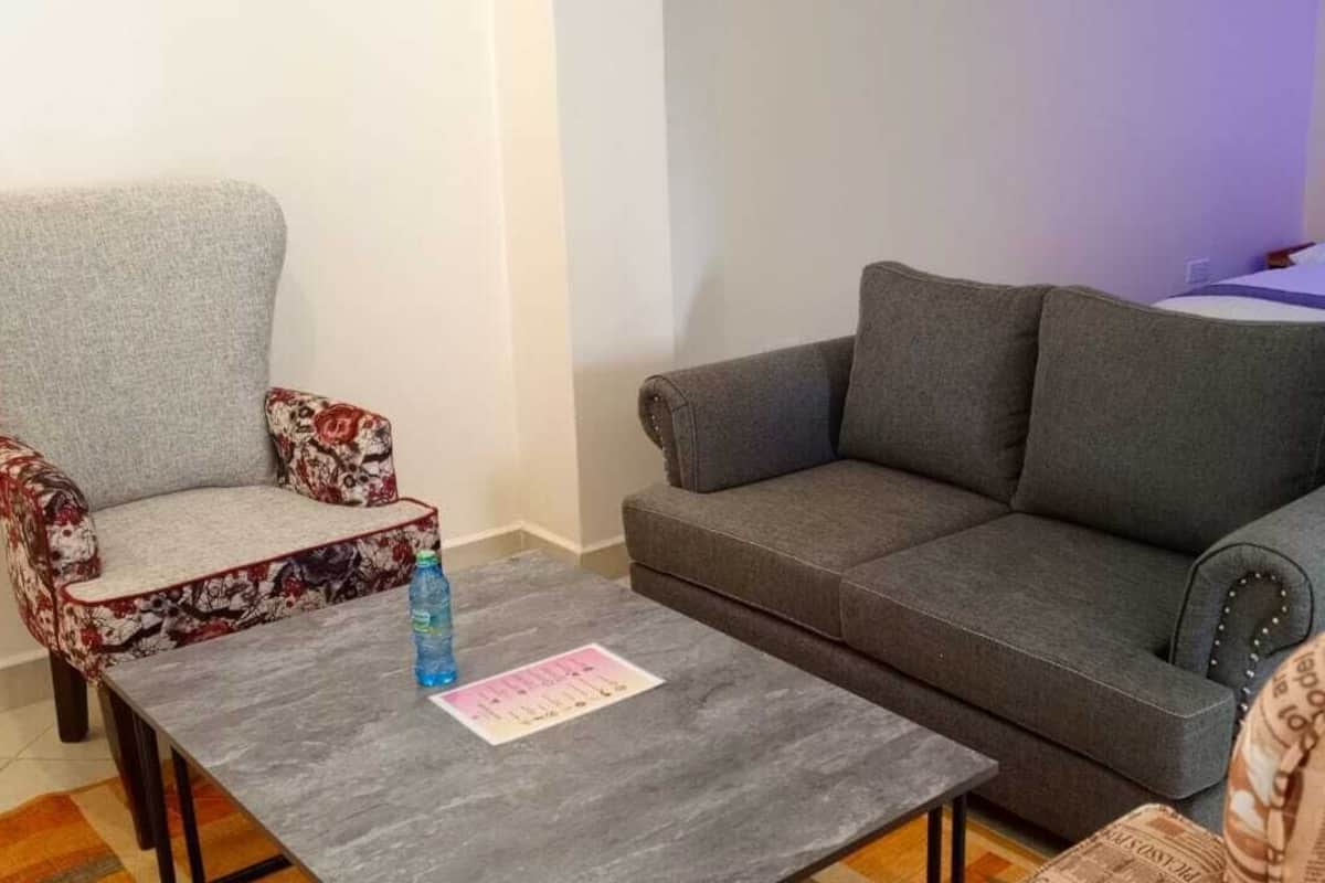 Quarto de hotel com um sofá cinzento, uma poltrona estampada, uma pequena mesa com uma garrafa de água e uma cama com edredom branco e almofadas.