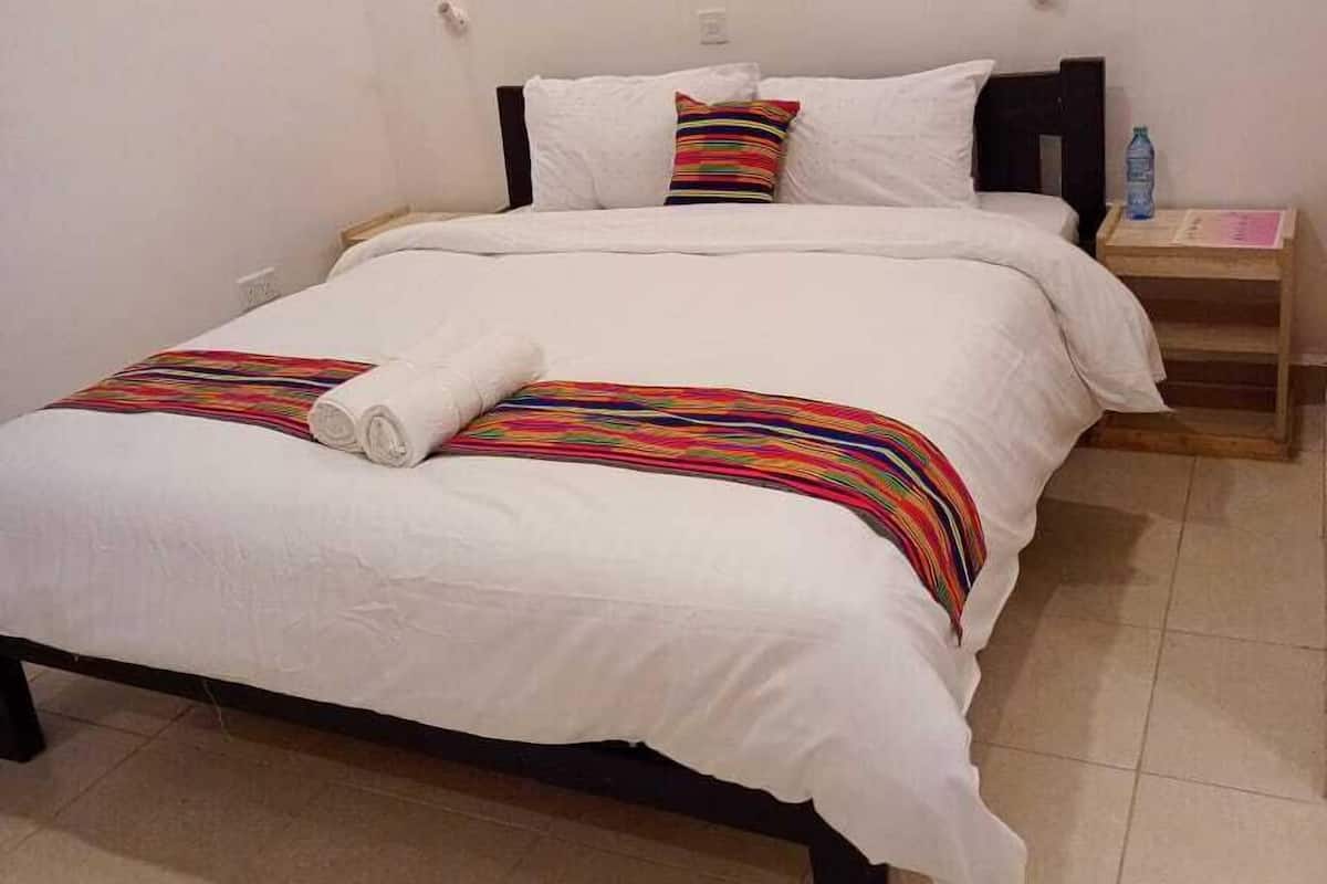 Uma cama bem arrumada com roupa de cama branca e um cobertor listrado colorido, uma pequena mesa de cabeceira com uma garrafa de água e uma parede branca lisa com uma luminária no teto.
