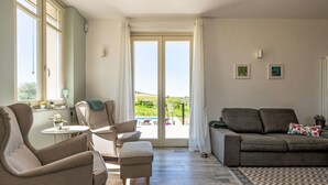 A spacious living room with a sofa, armchairs, and a small table. A large window offers a view of a green landscape.