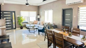 A modern living room with a sofa, dining table, chairs, ceiling fans, and large windows. - Your cool oasis home away from home. Breath taking views for relaxation  (Ocho Rios)