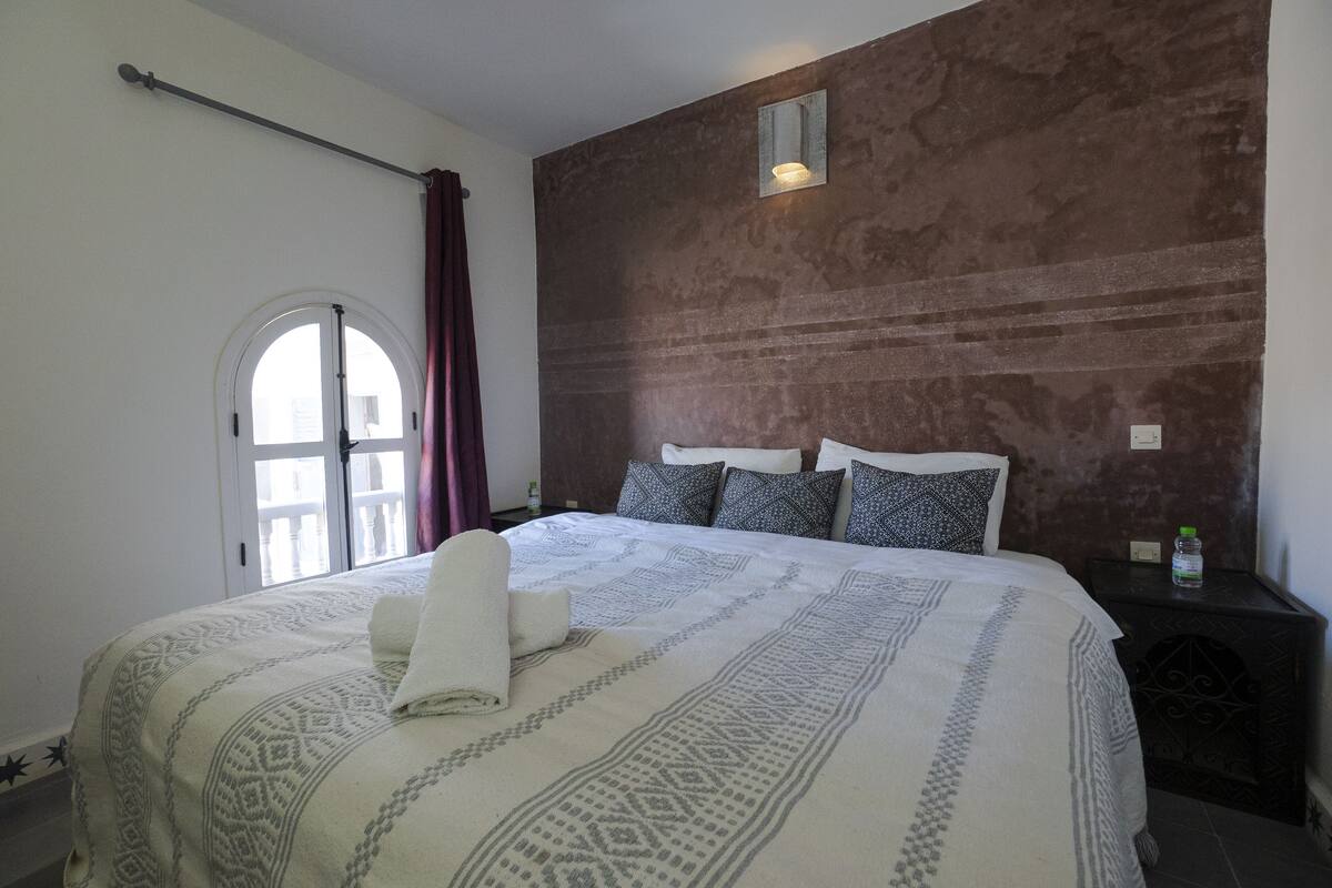 A bedroom with a large bed, a window with curtains, a wall-mounted light, and a nightstand with a bottle.