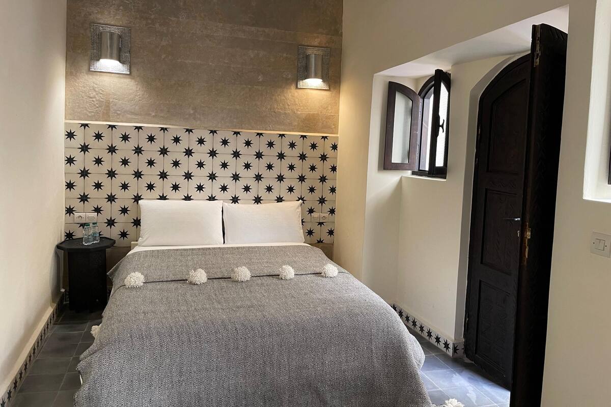 A bedroom with a star-patterned headboard, two wall-mounted lights, a bed with a patterned blanket, and a door with a window.