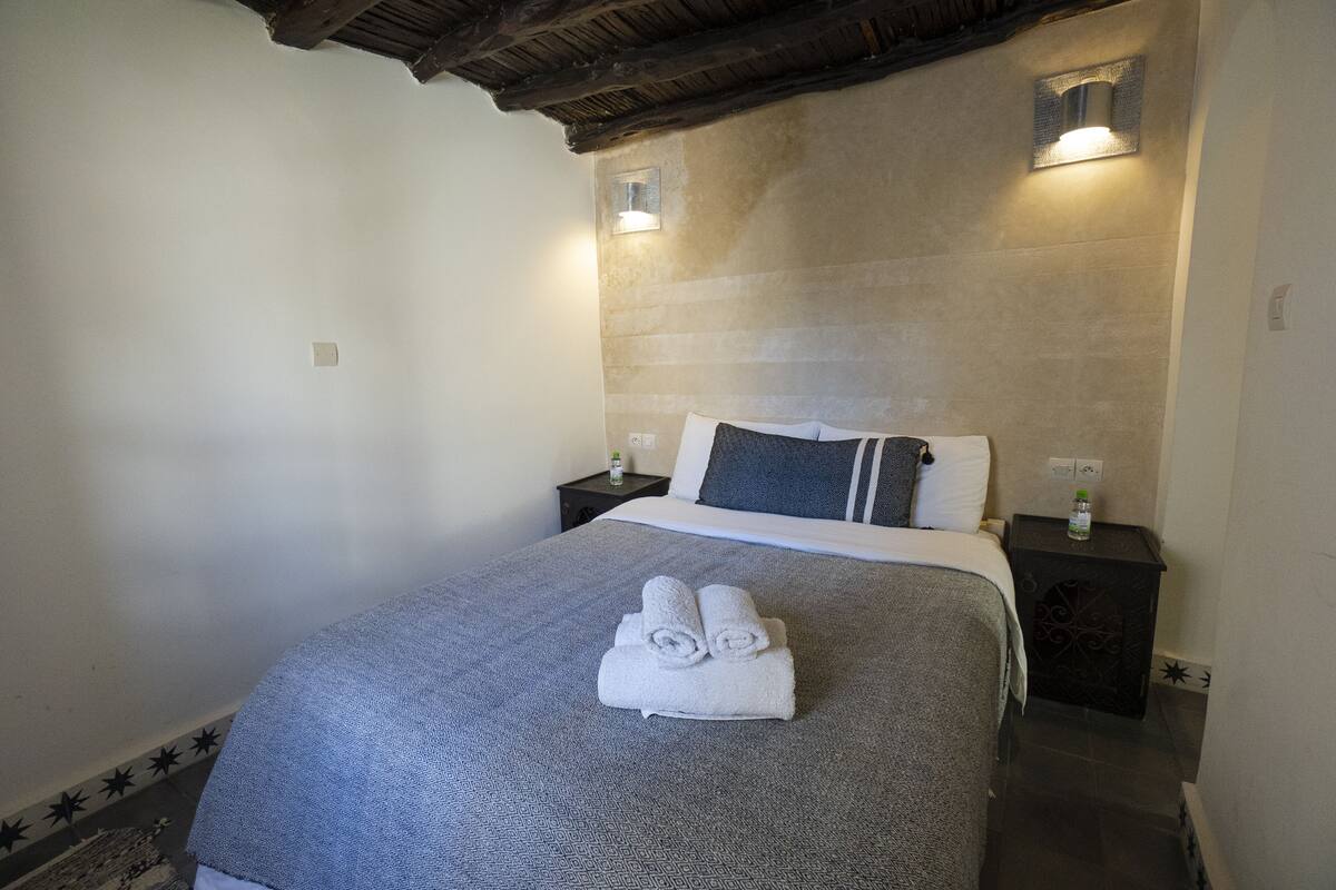 A bedroom with a bed, two bedside tables, a wall-mounted light, and a towel on the bed.