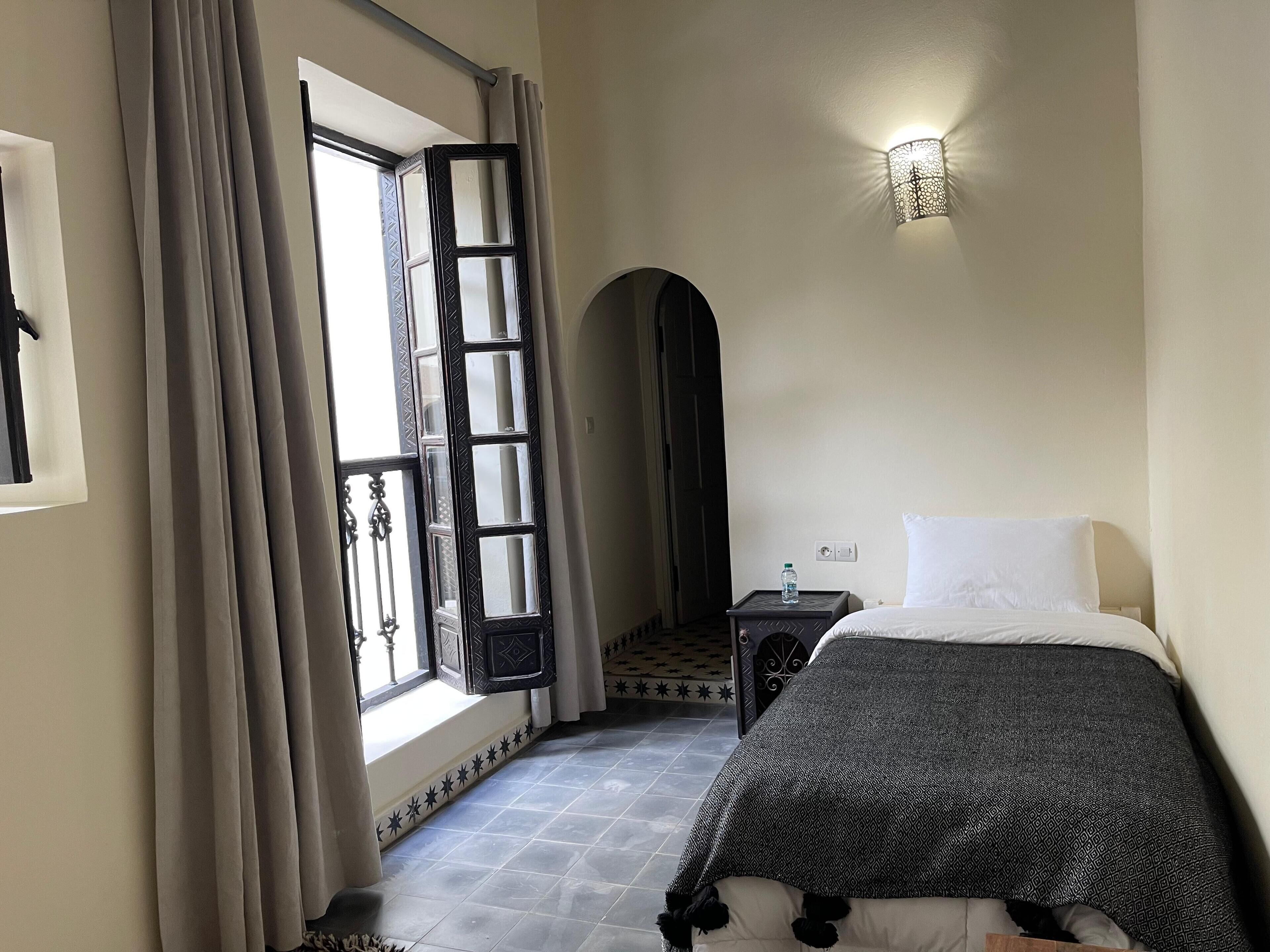 Ein Hotelzimmer mit Bett, Nachttisch, Wasserflasche, Wandleuchte und einem Fenster mit Vorhängen.