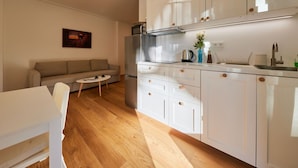 Une cuisine moderne avec des armoires blanches, un réfrigérateur en acier inoxydable et un four encastré. Le sol est en bois, et on y trouve une table à manger blanche avec des chaises, ainsi qu’une petite table basse ronde. Un canapé est placé dans l’espace salon, et un tableau encadré est accroché au mur.