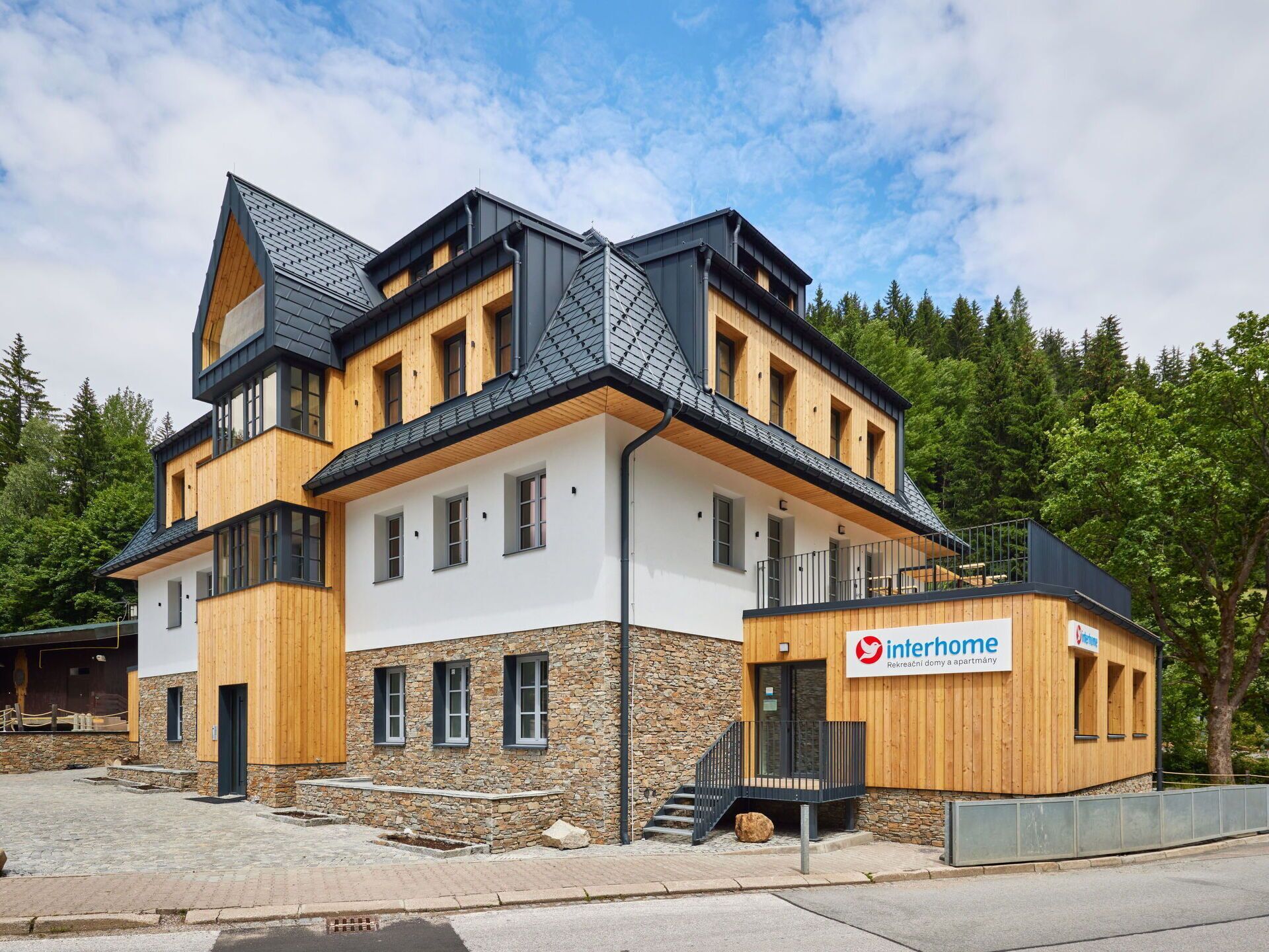 Un immeuble moderne à plusieurs étages, doté d’une combinaison de façades en bois et en pierre, d’un balcon et d’une petite annexe avec une enseigne.