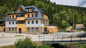 Un bâtiment moderne à deux étages, doté d’une façade en pierre et de détails en bois, situé près d’une route et surplombant un ruisseau par un pont piétonnier.