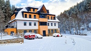 Un immeuble à plusieurs étages, avec une station de ski en toile de fond, dont la façade combine des éléments en bois et des surfaces blanches, et qui se dresse au milieu d’un paysage enneigé.
