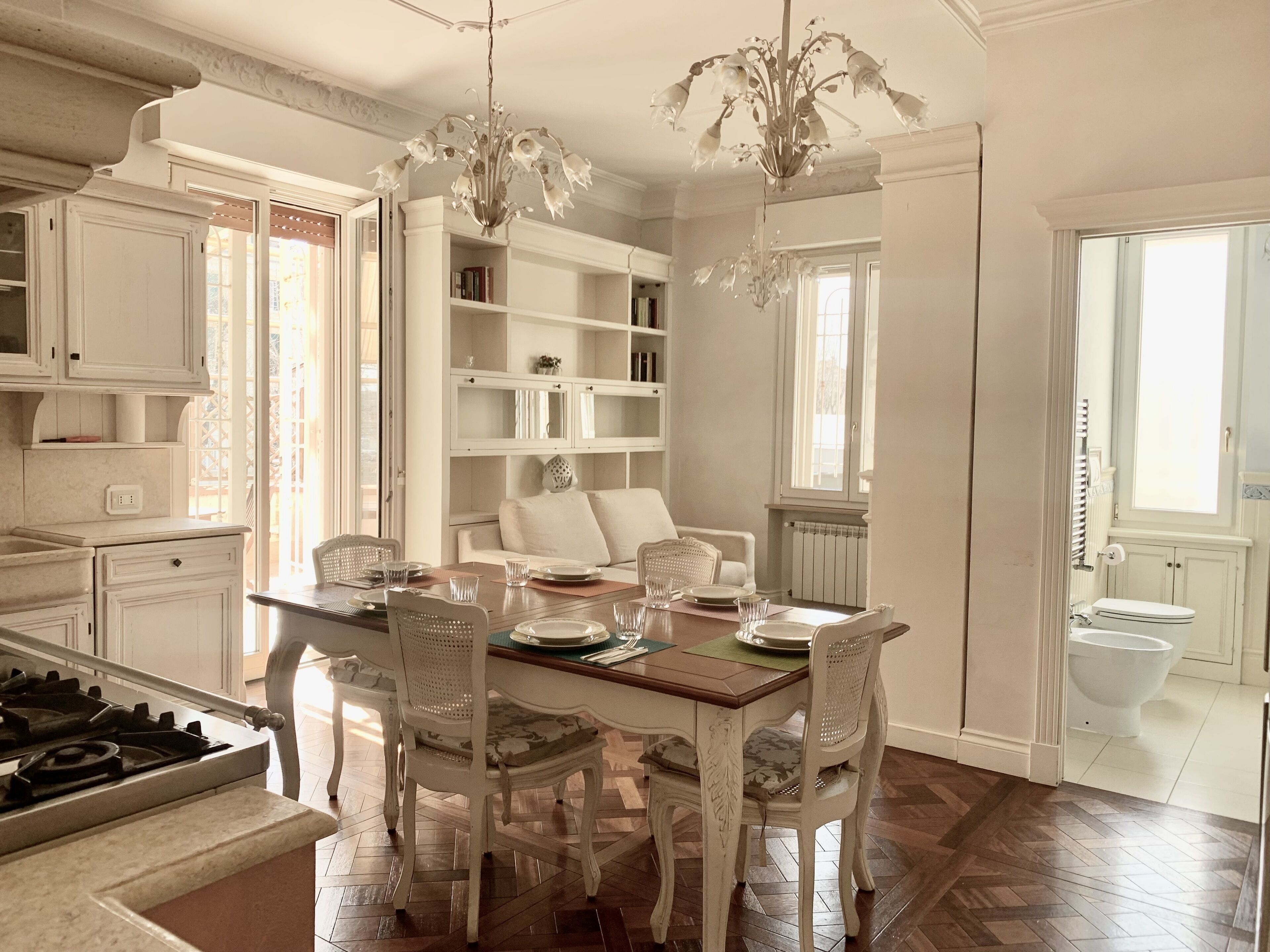 A dining area with a wooden table set for four, surrounded by white chairs. A chandelier hangs above, and there is a bookshelf with books and decorative items.