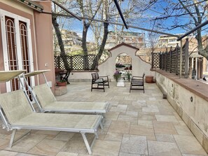 Eine Terrasse im Freien mit Metallstühlen und einem Tisch, umgeben von einer niedrigen Mauer und Bäumen.