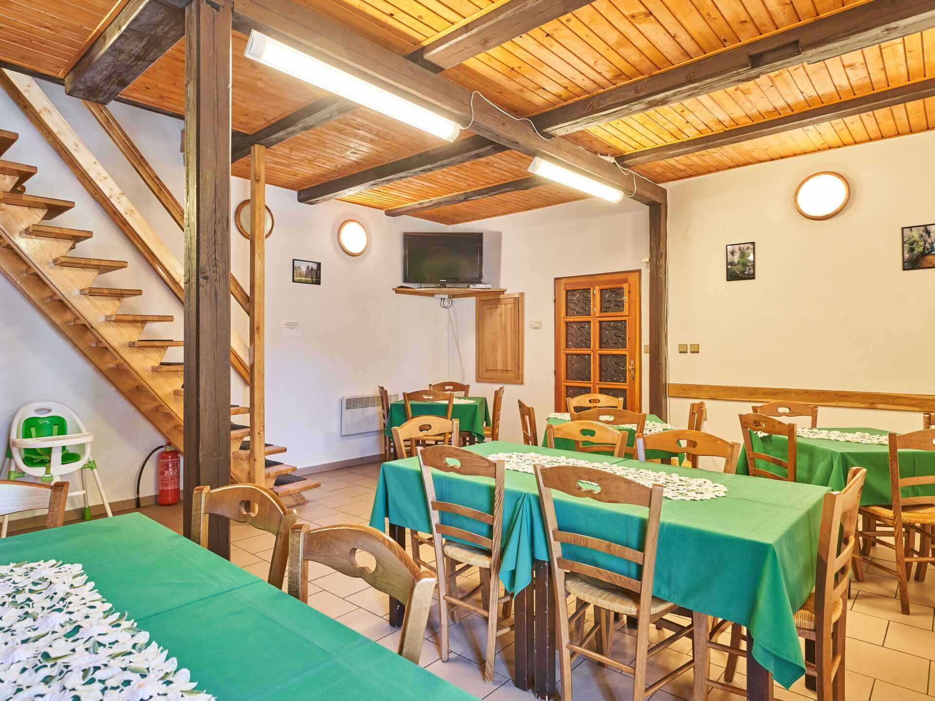 A dining area with wooden tables and chairs, a staircase, and a television on the wall.