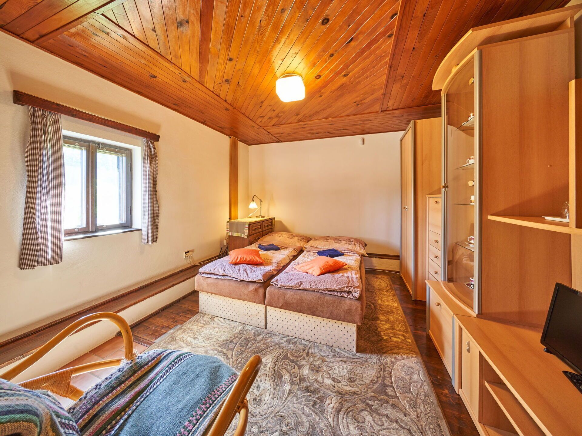 Una habitación acogedora con techo de madera, dos camas con ropa de cama estampada, una mesita de noche con lámpara, un armario y una ventana con cortinas.
