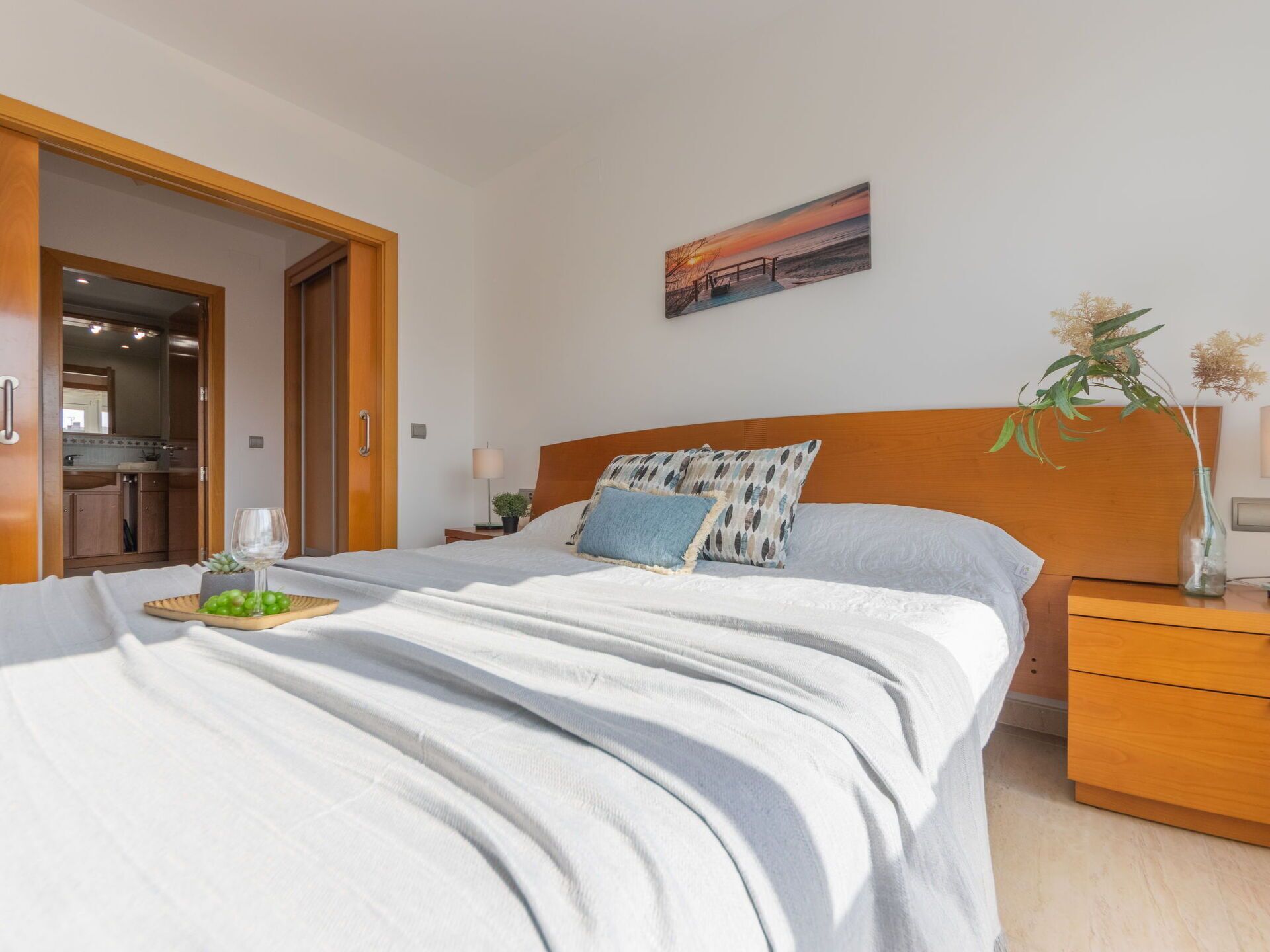 Un dormitorio con cama, mesita de noche, cabecera de madera, un cuadro colgado en la pared y una puerta que da al baño.
