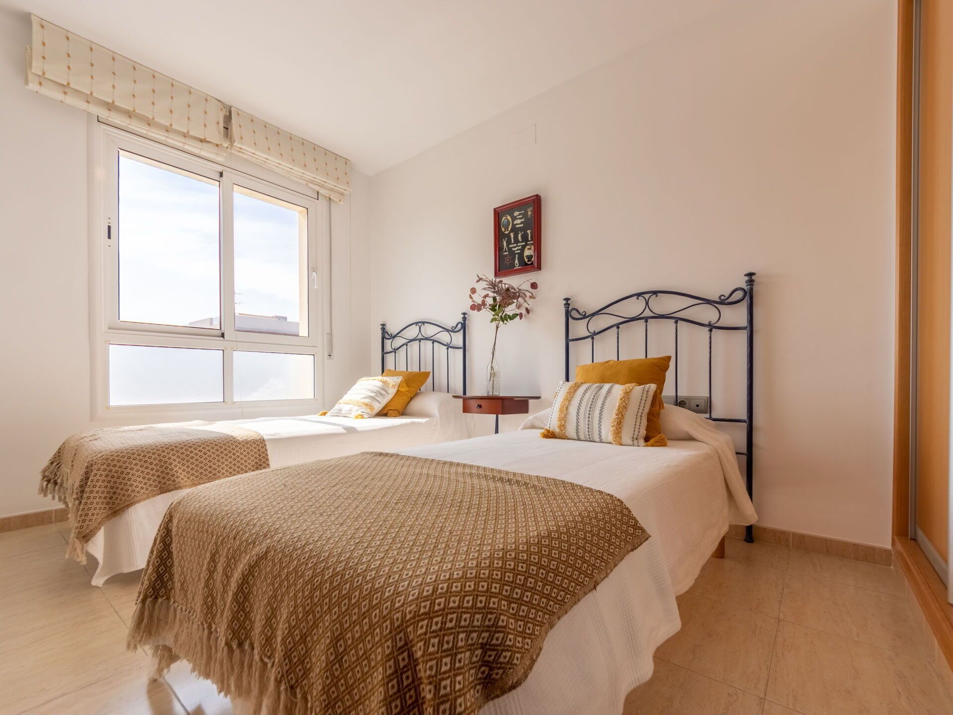 Un dormitorio con dos camas, una ventana con persianas, una mesita de noche con un florero y un reloj de pared.