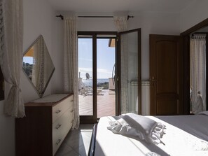 A bedroom with a bed, a dresser, a mirror, and a sliding glass door leading to an outdoor area with a view of the sea.