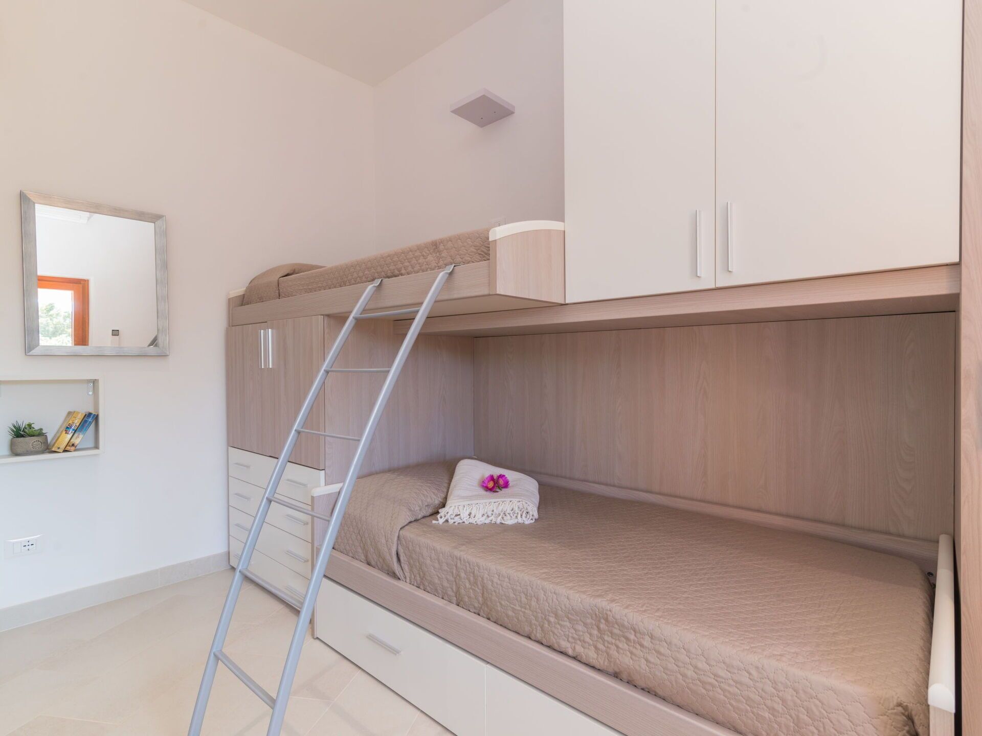 A bunk bed with a ladder, a small shelf with books and a plant, and a mirror on the wall.