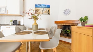 A modern kitchen with white cabinets, a dining table with four chairs, a vase with flowers, a clock, and a painting of a beach scene.
