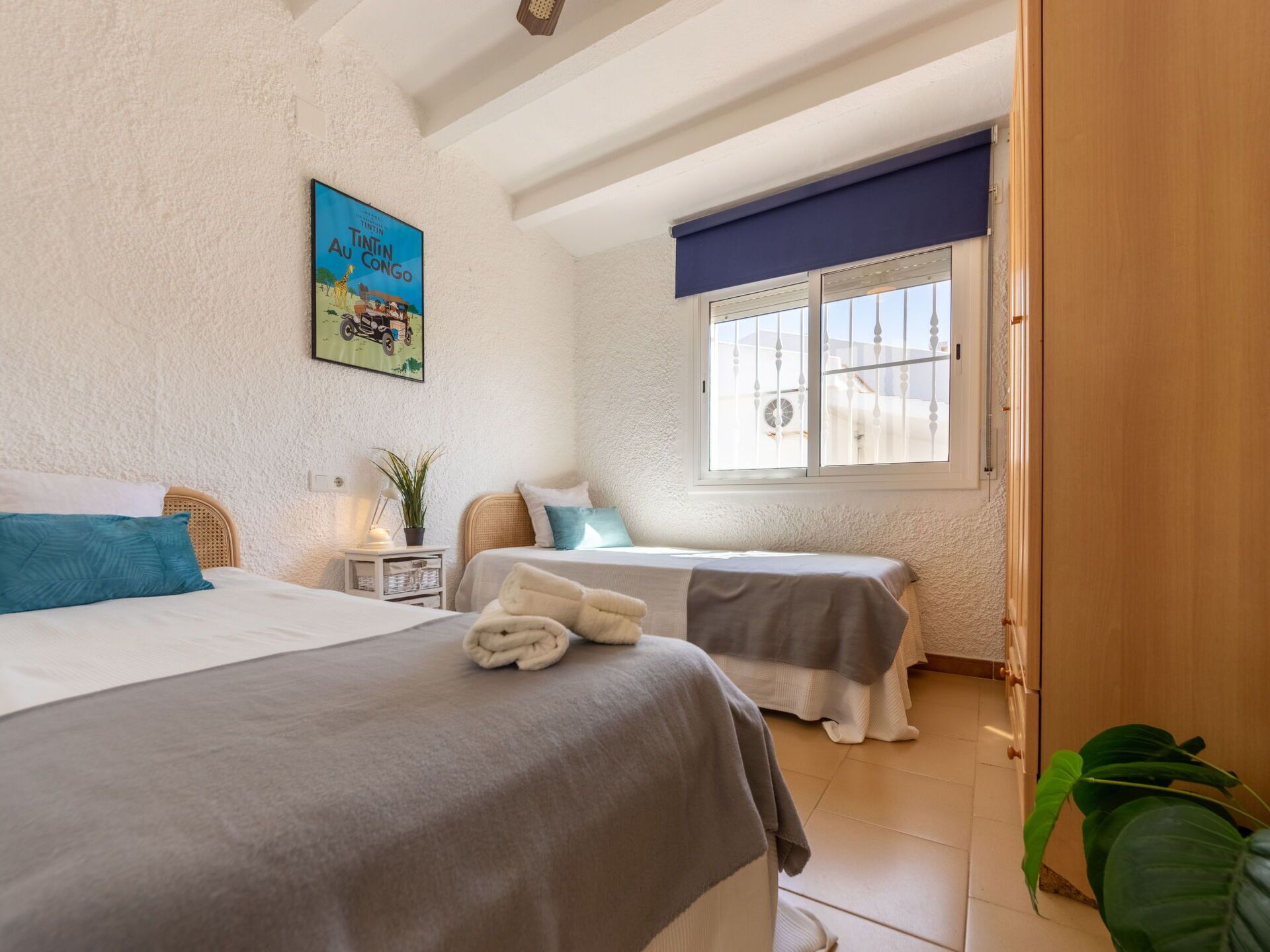 A bedroom with two beds, a window with a view, a painting on the wall, and a plant.