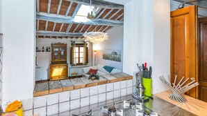 Une cuisine avec un mur recouvert de carreaux, des poutres apparentes, un luminaire suspendu, un meuble en bois et une fenêtre avec des rideaux.